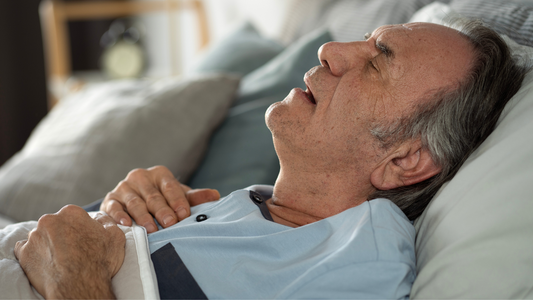 An elderly man sleeping on his back with his mouth open, wearing a light blue pajama top, lying in bed with his hands resting on his chest.